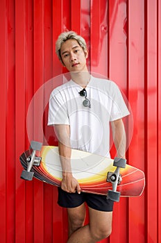 Young asian man in white T-shirt and sunglass holding the surfskate board in hip position in front of red galvanized steel sheet