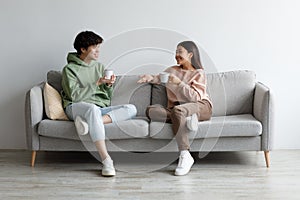 Young Asian man and woman drinking coffee and talking while sitting on couch at home