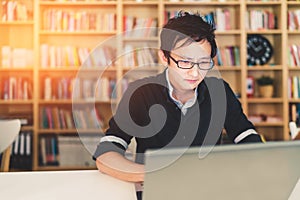 Young Asian man working on laptop at home office or library with serious face, bookshelf with clock blur background with clock