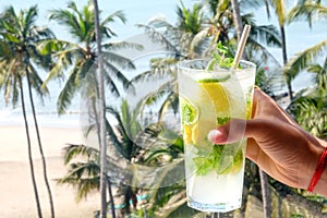 A young Asian mans hand holding a virgin mojito cocktail with palm trees the sea and a sandy beach in the background