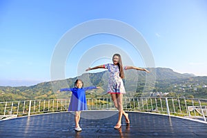 Young Asian mom and daughter feeling free with arms wide open at beautiful trees and mountains on blue sky