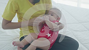 Young asian mother embracing and feeding little baby girl with bottle of milk on bed at home.