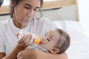 Young asian mother embracing and feeding little baby girl with bottle of milk at home.