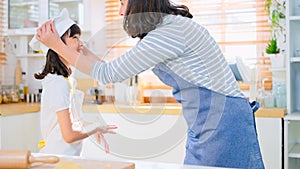 Young Asian mother help daughter wear apron and chef hat, prepare to learn bake pastry in kitchen at home