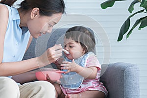 Young Asian mother holding spoon feeding child, Caucasian Cute 7 month newborn baby girl eating blend mashed food, sitting on sofa