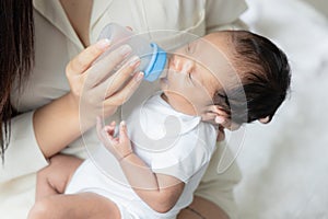 Young Asian mother hugging a cute newborn baby and feeding milk form bottle in white bedroom. Duty of the wife to raise children.