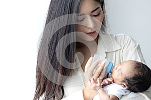 Young Asian mother hugging a cute newborn baby and feeding milk form bottle in white bedroom. Mother and child concept