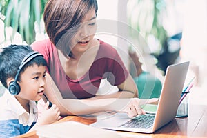 Young asian mother and son using laptop computer for study and learning together at home, boy wearing headphone.