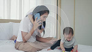 Young asian mother talking phone and playing with baby girl together on the bed, mom and daughter.