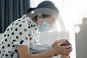 Young Asian mother wearing protective face mask playing with infant baby boy on bed in bedroom. Cheerful Mom with her Newborn