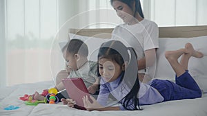 Young asian mother working at home online to internet with laptop computer and daughter looking tablet on bed.