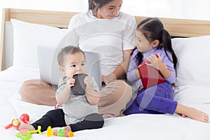 Young asian mother working at home online to internet with laptop computer and daughter looking tablet on bed at bedroom, business