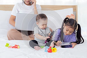 Young asian mother working at home online to internet with laptop computer and talking phone and daughter looking tablet.