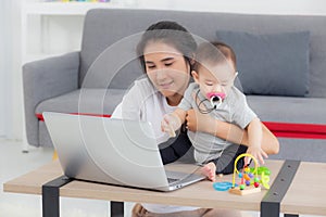Young asian mother working on laptop computer while care daughter girl at home.