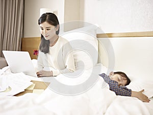 Young asian mother working on bed with sleeping daughter by side