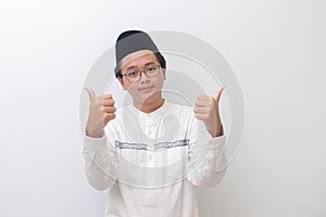 young Asian muslim man smiling and looking at camera, making thumbs up hand gesture. Isolated image on white