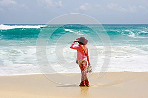 A young Asian muslim woman wearing hat, hijab and pink dress holding sandals standing on tropical beach looking at horizon