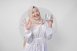 Young Asian Muslim woman in white dress and hijab holding and drinking cup of fresh water while making an okay, good, nice sign