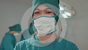 Young Asian nurse woman looking at camera and smiling after performing invasive surgery on patient in hospital operating room.