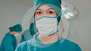 Young Asian nurse woman looking at camera and smiling after performing invasive surgery on patient in hospital operating room.