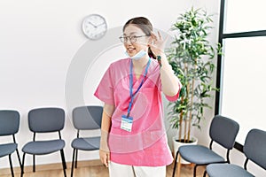 Young asian nurse woman at medical waiting room smiling with hand over ear listening an hearing to rumor or gossip