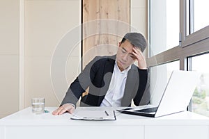 Young asian office worker suffering from headache at work in the office drinks medicine pill with water. Sick man in a suit