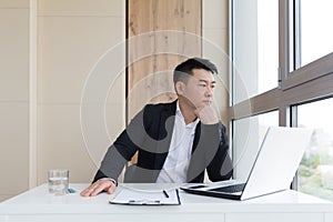 Young asian office worker suffering from headache at work in the office drinks medicine pill with water. Sick man in a suit