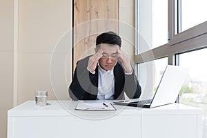 Young asian office worker suffering from headache at work in the office drinks medicine pill with water. Sick man in a suit