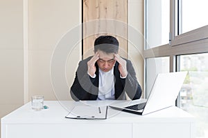 Young asian office worker suffering from headache at work in the office drinks medicine pill with water. Sick man in a suit