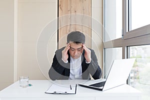 Young asian office worker suffering from headache at work in the office drinks medicine pill with water. Sick man in a suit