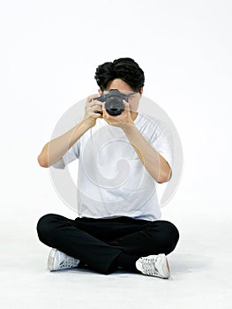 Young asian photographer in white t-shirt  looks through the viewfinder while sitting in a white photography scene. The atmosphere