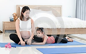 Young Asian pregnant woman and her first little kid daughter doing yoga, fitness exercises and stretching after workout