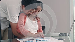 Young Asian Pregnant woman using laptop records of income and expenses at home. Dad touch his wife belly while record budget, tax
