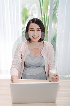 Young Asian pregnant woman working from home using computer and drinking milk for good health of her baby