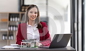 Young Asian real estate agent worker working with laptop at table in office and small house beside it.