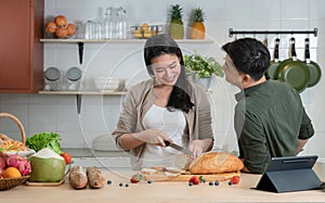 Young Asian romantic couple is cooking in the kitchen. Happy beautiful woman making healthy food like bread, fruits and salad