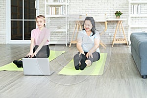 Young Asian and Senior woman sit on mat ready to exercising at home by watching video follow trainer online on laptop computer.