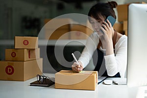 Young Asian small business owner working at home office, taking note on purchase orders. Online marketing packaging