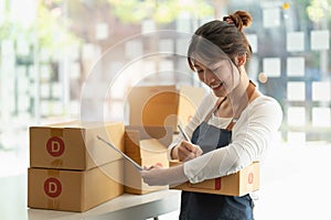 Young Asian small business owner working at home office, taking note on purchase orders. Online marketing packaging