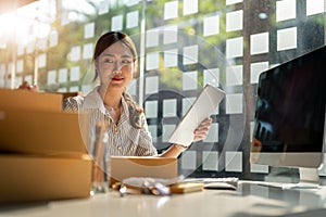 Young Asian small business owner working at home office, taking note on purchase orders. Online marketing packaging