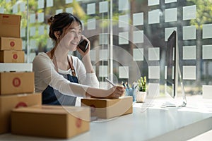 Young Asian small business owner working at home office, taking note on purchase orders. Online marketing packaging