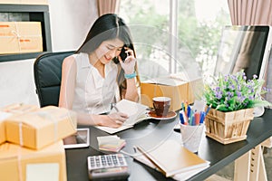 Young Asian small business owner working at home office, using mobile phone and taking note on purchase orders photo