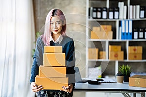 Young Asian SMEs woman holding parcels and looking to camera, standing among several boxes in modern office