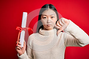 Young asian student woman holding graduate diploma over red  background with angry face, negative sign showing dislike