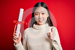 Young asian student woman holding graduate diploma over red isolated background happy with big smile doing ok sign, thumb up with