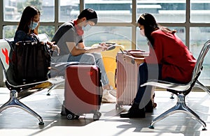 Young Asian tourists wearing casual dress and face masks with their luggage sitting in the airport terminal looking on smartphone