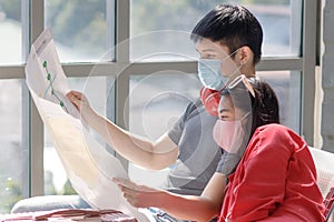 Young Asian travelers wearing casual dresses and face masks with their luggage sitting in the airport terminal looking at