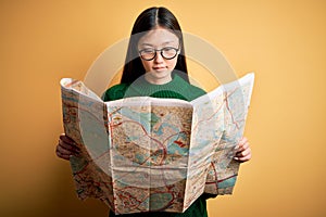 Young asian turist woman looking at city tourist map on a trip over yellow background with a confident expression on smart face