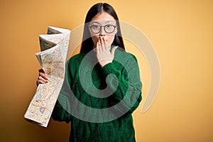 Young asian turist woman looking at city tourist map on a trip over yellow background cover mouth with hand shocked with shame for