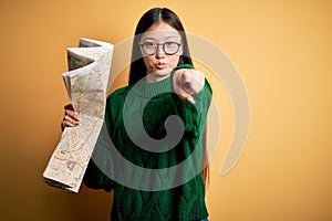 Young asian turist woman looking at city tourist map on a trip over yellow background pointing with finger to the camera and to